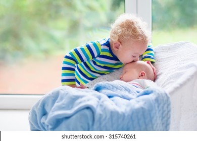 Cute little boy kissing his newborn brother. Toddler kid meeting new born sibling. Infant sleeping in white bouncer under a blanket. Kids playing and bonding. Children with small age difference.