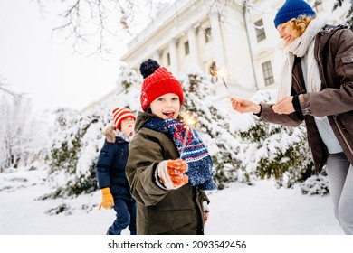 Cute little boy in knitted red hat holding fireworks with his family on background in a city park. Cute blonde child with dream.Happy kid enjoy the fire sparks. Holidays winter snow childhood concept.