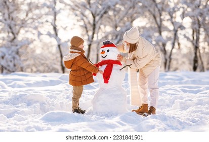 Cute little boy making snowman with mom in winter park. Happy young family mother and son in warm clothes building snow figure while playing and actively spending time on fresh frosty air in nature - Powered by Shutterstock