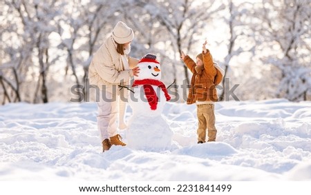 Cute little boy making snowman with mom in winter park. Happy young family mother and son in warm clothes building snow figure while playing and actively spending time on fresh frosty air in nature