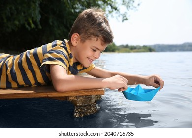 Cute little boy playing with paper boat on wooden pier near river