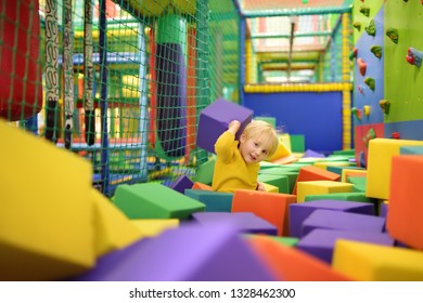 Cute Little Boy Plays With Soft Cubes In The Dry Pool In Play Center. Kid Playing On Indoor Playground In Foam Rubber Pit In Trampoline. Child Having Fun In Entertainment Center.