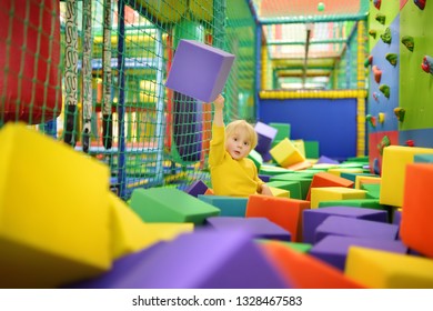 Cute Little Boy Plays With Soft Cubes In The Dry Pool In Play Center. Kid Playing On Indoor Playground In Foam Rubber Pit In Trampoline. Child Having Fun In Entertainment Center.