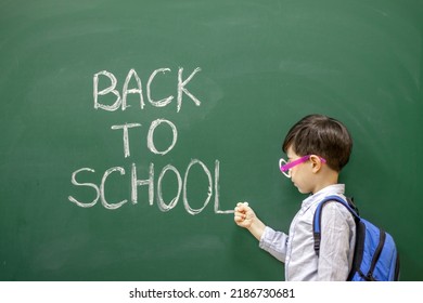 Cute Little Boy Preschooler Is Writing On Blackboard With White Chalk The Words Back To School.kid Is Wearing Funny Round Party Glasses With No Glass Inside.education,learning,classroom Interior