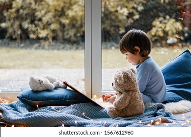 Cute little boy reading book with his teddy bear toy sitting cozy on pillows and knitted blanket near wet window with autumn garden at background. Cozy home. Winter holidays lifestyle. 