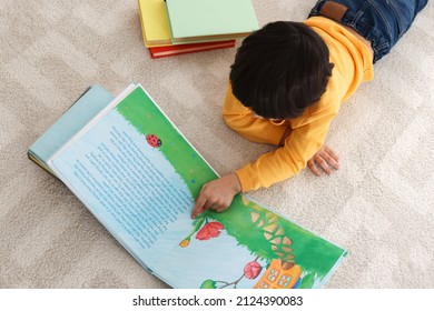 Cute little boy reading book on floor at home, above view