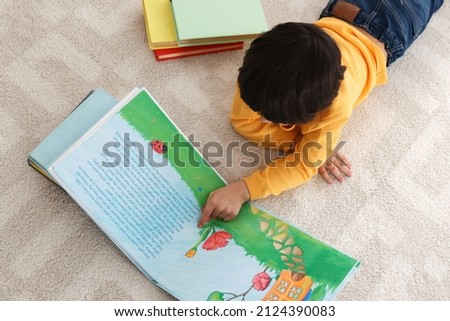 Cute little boy reading book on floor at home, above view