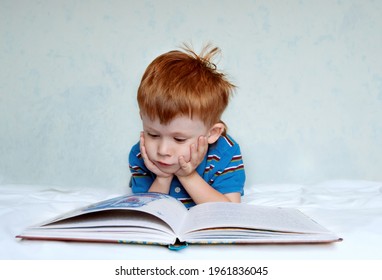 A Cute Little Boy With Red Hair And A Blue Shirt Is Lying On The Bed Reading A Book