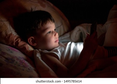 Cute Little Boy Sitting In The Dark And Watching Cartoons On Mobile. Smiling Toddler Laying On Bed And Looking At Smartphone. Light Reflection On Kid Face From Phone.