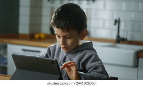 Cute little boy sitting in the kitchen playing mobile game on digital tablet device. Child surfing internet, play game at home. Children tech addiction concept - Powered by Shutterstock