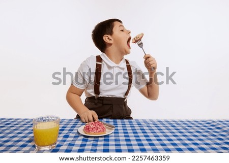 Cute little boy sitting at table covered plaid table cloth and tasting donut isolated on white background. Bored. Concept of emotions, retro style, fashion, 60s, 70s, family. Copy space for ad