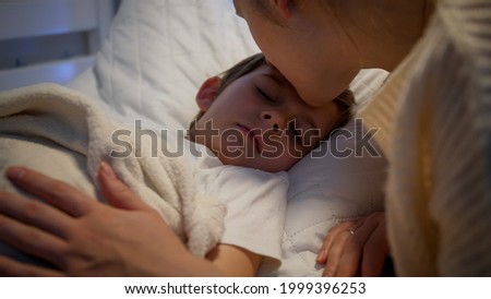 Cute little boy sleeping while caring mother kissing him and adjusting blanket. Concept of parenting and family happiness.