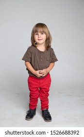 A cute little boy smiles affably and looks at the camera. Very long blond hair and thick bangs. Cute hairstyle in a child. He is standing in a studio in red pants and a brown t-shirt. Grey background