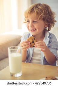 Cute Little Boy Is Smiling, Drinking Milk And Eating Cookies While Sitting On Couch At Home