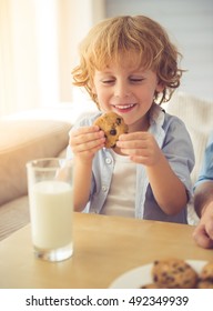 Cute Little Boy Is Smiling, Drinking Milk And Eating Cookies While Sitting On Couch At Home