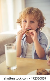 Cute Little Boy Is Smiling, Drinking Milk And Eating Cookies While Sitting On Couch At Home