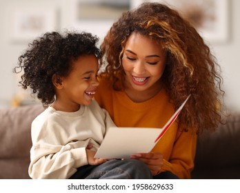 Cute little boy son congratulating his mom happy mixed race woman and Mothers day  giving her handmade greeting postcard and red heart while sitting together sofa at home  Family holidays concept