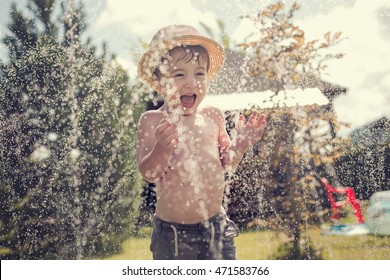 Cute little boy in straw hat is laughing and having fun running under water spraying hose. Image with selective focus and toning