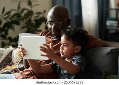 Cute little boy with tablet and his father sitting on couch in front of camera in living room and watching online video or cartoons - Powered by Shutterstock