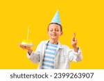 Cute little boy with tasty cake making Birthday wish on yellow background