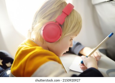 Cute Little Boy Traveling By An Airplane. Child Using Player To Listen A Music Or Audiobook During The Flight And Drawing Pictures. Entertainment For Family With Kids On A Board Of Plane