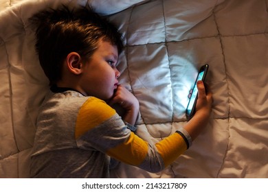 Cute little boy using a smartphone. Kid playing with mobile phone, lying on a bed. Freetime. Technology and internet concept. Smiling toddler laying on bed and looking at smartphone. Light reflectio