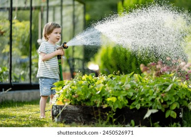 Cute little boy watering flower beds in the garden at summer day. Child using garden hose to water vegetables. Kid helping with everyday chores. Mommy's little helper. - Powered by Shutterstock