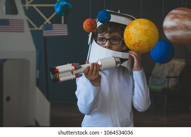 Cute little boy wearing cardboard astronaut helmet flying toy rocket through planets, cardboard spaceship rocket in the background - Shutterstock ID 1064534036