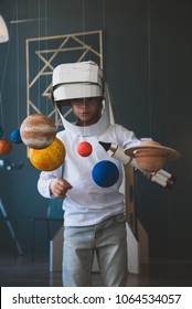 Cute little boy wearing cardboard astronaut helmet flying toy rocket through planets, cardboard spaceship rocket in the background - Shutterstock ID 1064534057