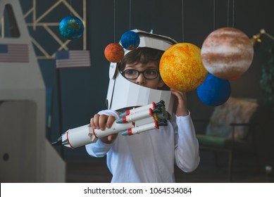Cute little boy wearing cardboard astronaut helmet flying toy rocket through planets, cardboard spaceship rocket in the background - Shutterstock ID 1064534084