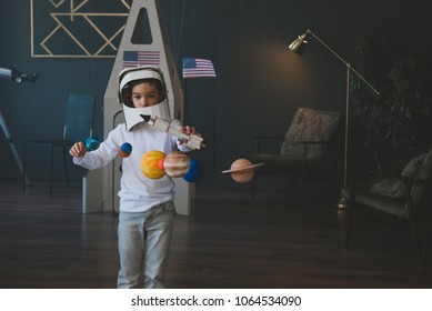 Cute little boy wearing cardboard astronaut helmet flying toy rocket through planets, cardboard spaceship rocket in the background - Shutterstock ID 1064534090