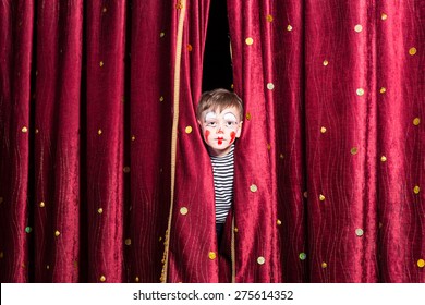 Cute Little Boy Wearing Colorful Red Face Paint And A Costume Peering Out From Between The Curtains On Stage As He Waits For A Pantomime To Begin