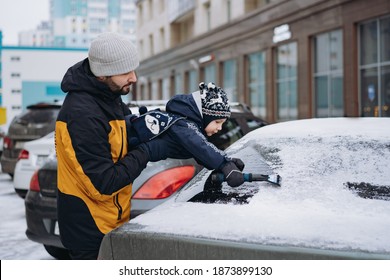 Cute Little Boy Wearing Scandinavian Knit Hat And Scarf Helping To Brush Snow From A Car On Winter Day  Near Apartment Building. Father Is Lifting Son To Reach The Window. Image With Selective Focus