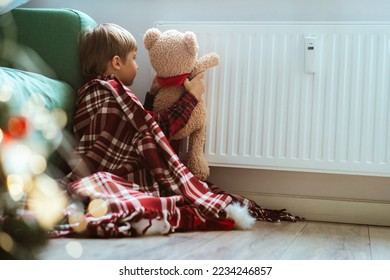 cute little boy wrapped id plaid sitting by heater warming teddy bear paws. Christmas time. Cold at home, Energy crisis. Fuel poverty - Shutterstock ID 2234246857