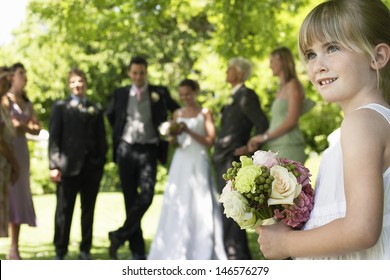 Cute little bridesmaid holding bouquet in lawn with guests and wedding couple in background