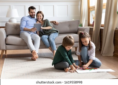 Cute Little Brother And Sister Sit On Floor Drawing In Album, Young Parents Relax On Comfortable Couch In Living Room Using Tablet, Young Caucasian Family Rest Together Enjoy Leisure Weekend At Home