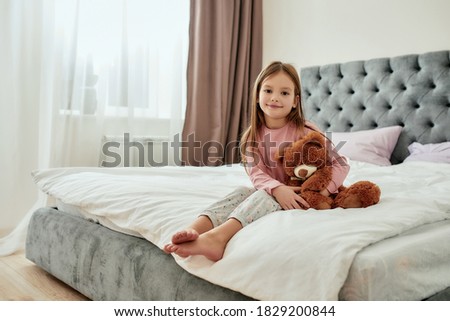 A cute little browneyed girl sitting barefoot on a huge bed holding her teddybear with her feet crossed wearing pyjamas in a bright spacy bedroom. Happy childhood concept