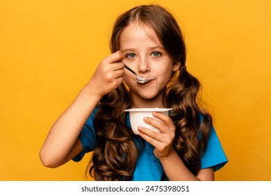 Cute little brunette girl in blue t-shirt eating tasty yogurt on light yellow background. Healthy nutrition concept. Copy space. - Powered by Shutterstock