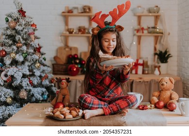 Cute Little Brunette Girl In Checkered Red Pajamas With Deer Horns On Her Head Is Eating A Christmas Cake In A Beautifully Decorated Kitchen. Winter New Year's Tale. Family Tea Party. Hygge Time