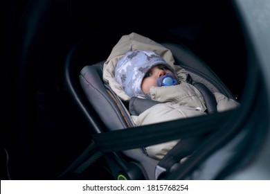 Cute Little Caucasian Dummy Sucking Baby Boy Lying In Car Child Seat Fastened With Belt Wearing Winter Overall And Hat. Image With Selective Focus.