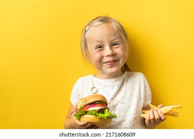 Cute Little Caucasian Girl With Blonde Hair Enjoying Burger On A Yellow Background. Happy Kid Smiling And Eating Fast Food Burger