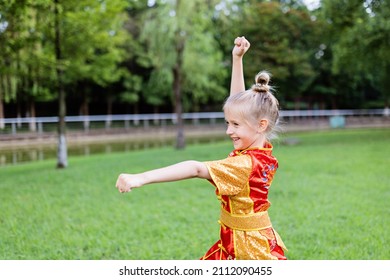 Cute Little Caucasian Girl Seven Years Old In Red Sport Wushu Uniform Exercising In Park At Summer Day. Lifestyle Portrait Of Kung Fu Fighter Child Athlete