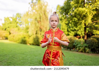 Cute Little Caucasian Girl Seven Years Old In Red Sport Wushu Uniform Exercising In Park At Summer Day. Lifestyle Portrait Of Kung Fu Fighter Child Athlete