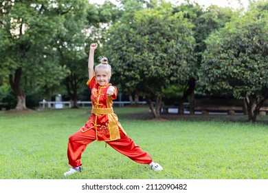 Cute Little Caucasian Girl Seven Years Old In Red Sport Wushu Uniform Exercising In Park At Summer Day. Lifestyle Portrait Of Kung Fu Fighter Child Athlete