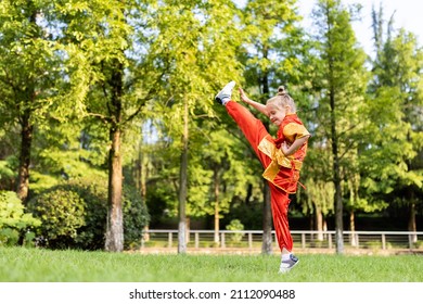 Cute Little Caucasian Girl Seven Years Old In Red Sport Wushu Uniform Exercising In Park At Summer Day. Lifestyle Portrait Of Kung Fu Fighter Child Athlete