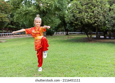 Cute Little Caucasian Girl Seven Years Old In Red Sport Wushu Uniform Exercising In Park At Summer Day. Lifestyle Portrait Of Kung Fu Fighter Child Athlete