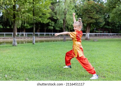 Cute Little Caucasian Girl Seven Years Old In Red Sport Wushu Uniform Exercising In Park At Summer Day. Lifestyle Portrait Of Kung Fu Fighter Child Athlete