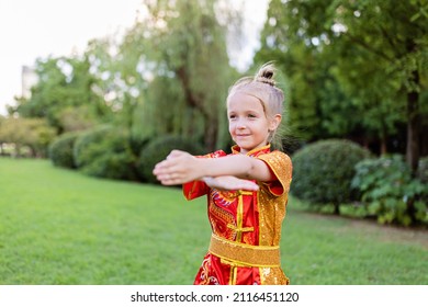 Cute Little Caucasian Girl Seven Years Old In Red Sport Wushu Uniform Exercising In Park At Summer Day. Lifestyle Portrait Of Kung Fu Fighter Child Athlete