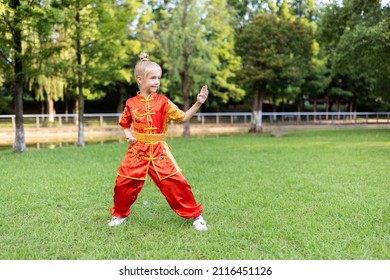 Cute Little Caucasian Girl Seven Years Old In Red Sport Wushu Uniform Exercising In Park At Summer Day. Lifestyle Portrait Of Kung Fu Fighter Child Athlete