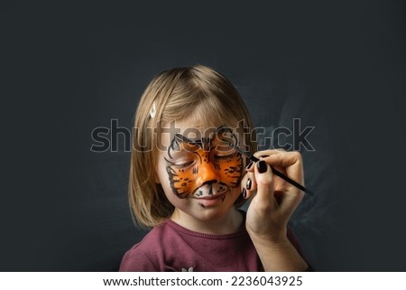 Cute little caucasian girl with tiger face painting on the black background. Close up portrait of little kid with face-painting. Year of a tiger. Happy smiling girl pretending she is a tiger